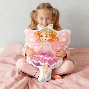 Op de afbeelding: Een vlindervormig kussen met een cartoonfee. De fee heeft oranje haar, een roze jurk en blauwe schoenen. Het kussen heeft roze vleugels en de naam "Sophia" op de voorkant. Een persoon in een roze pyjama houdt het kussen vast.
