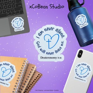 May include: A collection of stickers, a water bottle, a laptop, a phone, and notebooks are displayed on a purple background. The stickers feature a blue heart with a cross and the text "I am never alone. God will never leave me. Deuteronomy 31:6."