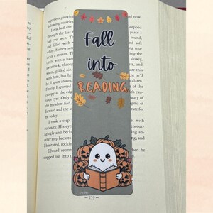 Puede incluir: Marcapáginas rectangular con fondo gris y las palabras "Fall into Reading" en negro y naranja. El marcapáginas presenta un fantasma lindo leyendo un libro, rodeado de calabazas. Hojas de otoño decoran la parte superior.