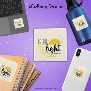 Pode incluir: Uma variedade de itens, incluindo um laptop, garrafa de água, telefone e cadernos, cada um adornado com um adesivo com o texto "BE THE light." O design inclui um sol amarelo e a referência bíblica Mateus 5:14.