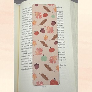 Pode incluir: Um marcador de livros retangular com fundo bege, adornado com um padrão repetido de folhas de outono, abóboras e bolotas em tons de laranja, castanho e verde. O marcador de livros está colocado num livro aberto. O número "259" é visível na parte inferior.