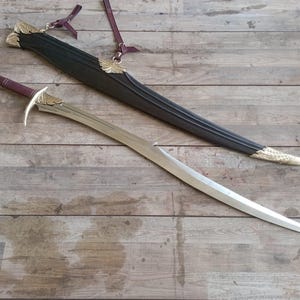 May include: A curved sword with a metallic blade, a maroon leather-wrapped handle, and gold-colored wing-shaped accents on the crossguard. It is accompanied by a black scabbard featuring matching gold-colored wing details and maroon leather straps.