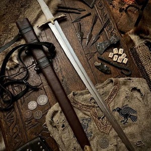 Peut inclure: Une longue épée en acier avec une poignée blanche et une garde dorée, accompagnée d'un fourreau en cuir marron avec une pointe en laiton. L'ensemble comprend plusieurs pointes de flèches en fer, des runes en bois, des pièces et une tunique.