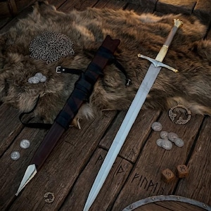 Può includere: Una lunga spada in acciaio con guardia e pomolo metallici, accompagnata da un fodero in pelle marrone scuro con cinghie nere. Il set è esposto insieme a una cotta di maglia, monete d'argento, una spilla in metallo e due dadi runici in legno.