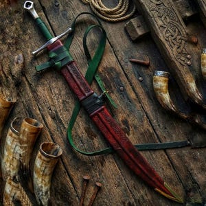 Può includere: Una spada in stile medievale con un'elsa e un fodero avvolti in pelle rossa e marrone. La spada presenta una guardia incrociata argentata e un cinturino in pelle verde. Lo sfondo include elementi in legno, chiodi e corna potorie.