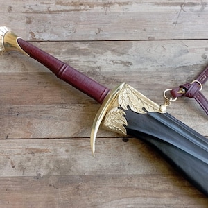 May include: A sword with a dark maroon leather-wrapped handle, a polished brass pommel, and a curved brass crossguard featuring leaf engravings. The black scabbard is accented with brass leaf details and includes a maroon leather strap attachment.