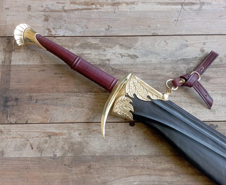May include: A sword with a dark maroon leather-wrapped handle, a polished brass pommel, and a curved brass crossguard featuring leaf engravings. The black scabbard is accented with brass leaf details and includes a maroon leather strap attachment.