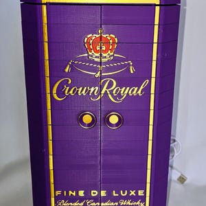 May include: A purple cabinet with gold trim and the Crown Royal logo. The cabinet has two doors with gold knobs and the words "Fine de Luxe Blended Canadian Whisky" are printed on the front.