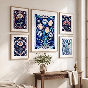 Op de afbeelding: Een galerijwand met vijf ingelijste prints met botanische illustraties van tulpen en klaprozen in tinten diepblauw, wit en zachtroze, weergegeven in lichte houten lijsten aan een effen muur boven een houten sidetable.