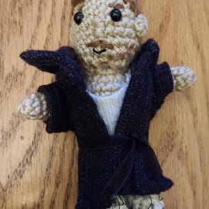 May include: Handmade crochet doll with a light brown head, dark eyes, and a small mustache. The doll wears a dark blue robe with a black belt and white shirt. The doll's feet are gray and white.