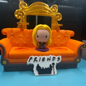 May include: An orange miniature couch with a gold back and a figurine of a blonde woman holding a guitar. A sign that says "FRIENDS" is in front of the couch. The couch has two cup holders and a gray base.