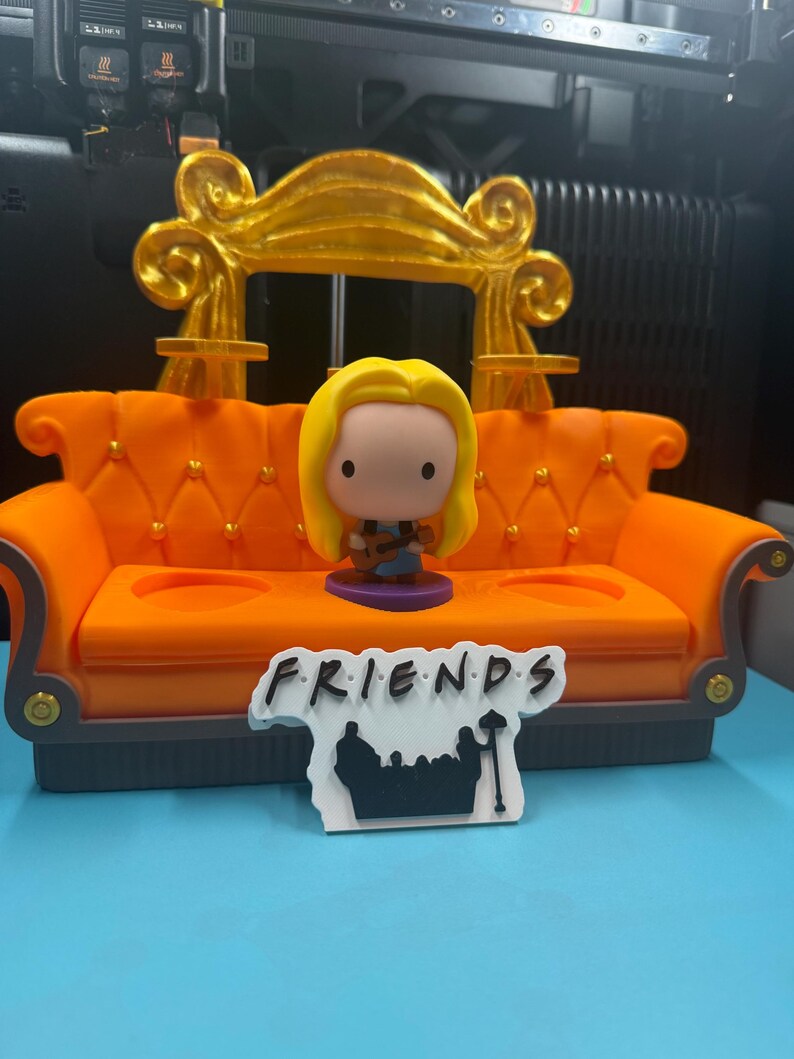 May include: An orange miniature couch with a gold back and a figurine of a blonde woman holding a guitar. A sign that says "FRIENDS" is in front of the couch. The couch has two cup holders and a gray base.