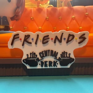 May include: A decorative sign featuring the word "FRIENDS" in a stylized font with colorful dots. Below, the sign reads "CENTRAL PERK" with coffee cup illustrations. The sign is set against an orange couch.