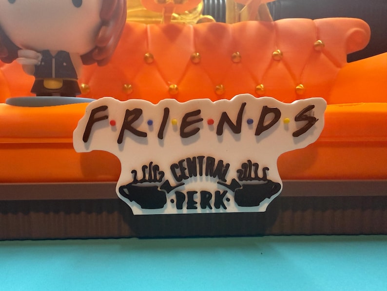 May include: A decorative sign featuring the word "FRIENDS" in a stylized font with colorful dots. Below, the sign reads "CENTRAL PERK" with coffee cup illustrations. The sign is set against an orange couch.