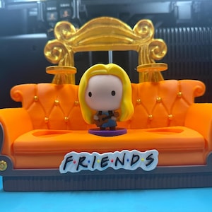 May include: A miniature orange couch with a gold-colored decorative element on the back. A figurine of a woman with blonde hair is standing in front of the couch. The word "FRIENDS" is written on the front of the couch.