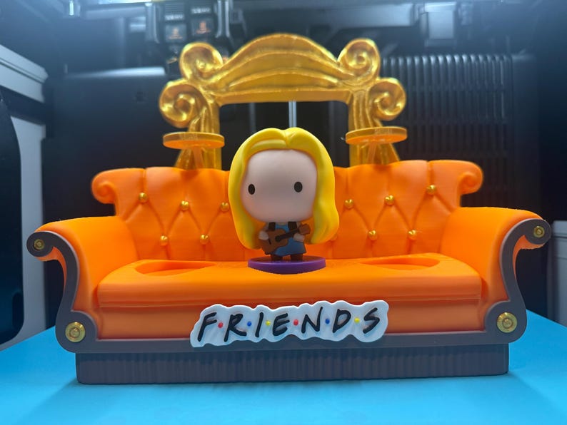 May include: A miniature orange couch with a gold-colored decorative element on the back. A figurine of a woman with blonde hair is standing in front of the couch. The word "FRIENDS" is written on the front of the couch.