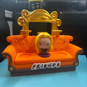 May include: An orange miniature couch with a gold-colored back and the word "FRIENDS" on the front. A Funko Pop figure of a character with blonde hair is seated on the couch. The couch has two cup holders and decorative gold accents.