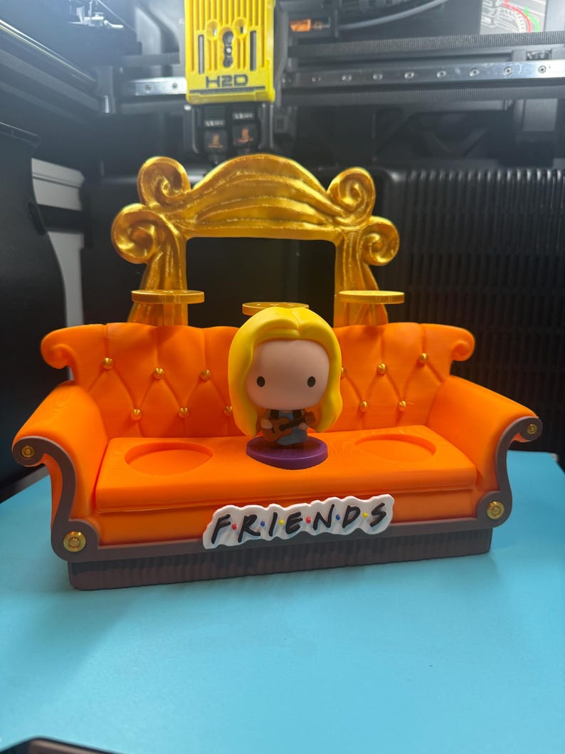 May include: An orange miniature couch with a gold-colored back and the word "FRIENDS" on the front. A Funko Pop figure of a character with blonde hair is seated on the couch. The couch has two cup holders and decorative gold accents.