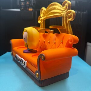 May include: A 3D-printed orange couch with a gold backrest, featuring a cartoon character figurine. The couch has a brown base and gold accents. The word "Friends" is displayed on the front of the couch.
