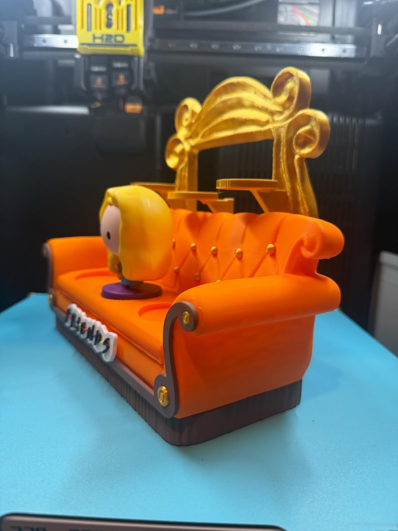 May include: A 3D-printed orange couch with a gold backrest, featuring a cartoon character figurine. The couch has a brown base and gold accents. The word "Friends" is displayed on the front of the couch.