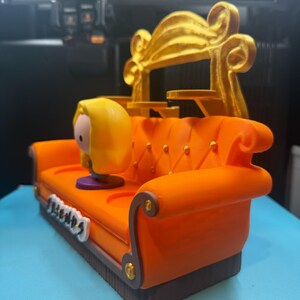 May include: An orange miniature couch with a gold back, featuring a cartoon character with yellow hair. The couch has the word "FRIENDS" on the front. The couch is a decorative item.