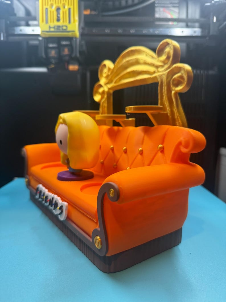 May include: An orange miniature couch with a gold back, featuring a cartoon character with yellow hair. The couch has the word "FRIENDS" on the front. The couch is a decorative item.