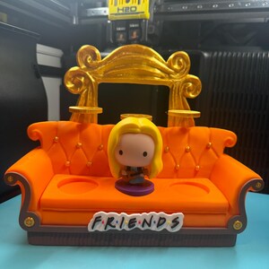 May include: Orange miniature couch with a gold-colored back and the word "FRIENDS" on the front. A small figurine of a character with blonde hair is sitting on the couch. The couch has two circular cutouts.