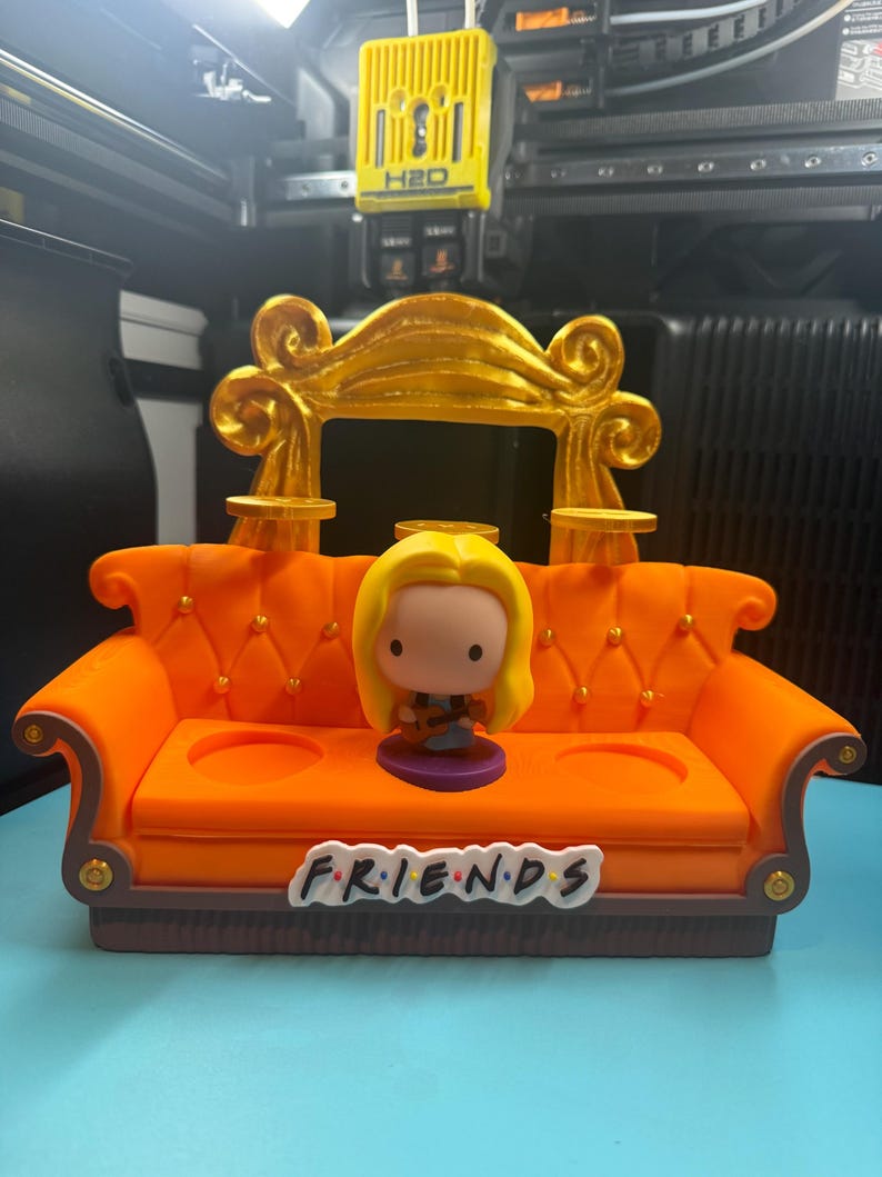 May include: Orange miniature couch with a gold-colored back and the word "FRIENDS" on the front. A small figurine of a character with blonde hair is sitting on the couch. The couch has two circular cutouts.
