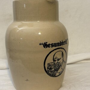 May include: A beige ceramic pitcher with a smooth, glossy finish. The front features a black circular graphic depicting a bearded person holding a mug, accompanied by the text "Gesundheit!" printed in a bold, black, gothic-style typeface.
