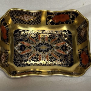 Puede incluir: Un plato rectangular de porcelana con bordes festoneados, que presenta una paleta de colores azul marino profundo y dorado. El interior muestra un intrincado patrón de medallón floral con acentos naranjas, rodeado por un borde de celosía y volutas doradas.
