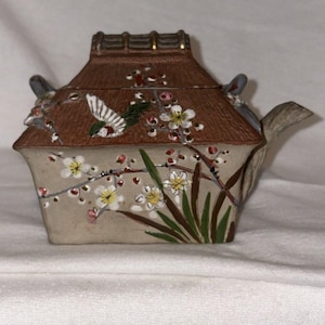 Peut inclure: Une théière en céramique avec un couvercle en forme de toit marron et un corps beige. La théière est décorée d'un oiseau, de fleurs blanches et de feuilles vertes. Le bec et la anse sont beige clair.