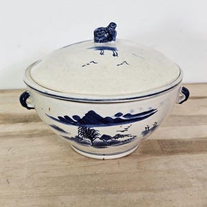 May include: A white and blue ceramic serving bowl with a lid. The bowl features a landscape design with mountains, trees, and birds. The lid has a small blue figurine on top. The bowl has two handles.