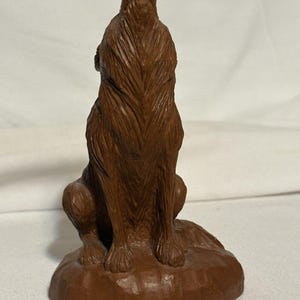 May include: A brown wooden sculpture of a wolf howling upwards. The wolf is sitting on a small, round base. The sculpture has detailed carved fur and a textured appearance. The wolf's head is tilted back, mouth open, in a howling pose.