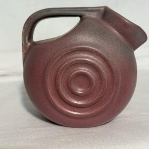Może przedstawiać: Okrągły ceramiczny dzbanek w matowym, malinowym odcieniu o teksturowanej powierzchni. Korpus posiada trzy koncentryczne, wytłoczone pierścienie na środku. Dzbanek ma zakrzywiony uchwyt przymocowany u góry oraz mały dzióbek po prawej stronie.