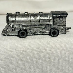 May include: A small, silver toy train with two black wheels on each side. The train has a detailed design, including a smokestack, windows, and a front cowcatcher. The toy is likely made of metal.