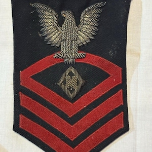Pode incluir: Um emblema de patente militar em tecido preto com uma águia dourada bordada no topo, posicionada sobre um design de chevron vermelho. No centro do chevron vermelho há uma forma de diamante contendo a letra M bordada em fio dourado.