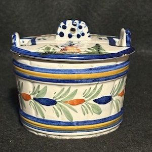 Könnte beinhalten: Ein Keramikbehälter mit Deckel, handbemalt mit einem dekorativen Design. Der Behälter ist weiß mit blauen, gelben und orangenen Akzenten. Der Deckel hat einen Griff mit blauen Tupfen. Der Körper ist mit einem Blatt- und Beerenmuster verziert.