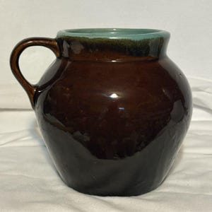 May include: A rounded ceramic pitcher with a glossy dark brown glaze covering the body and handle. The interior rim and the top edge of the vessel feature a contrasting light blue-green glaze that drips slightly down into the brown finish.