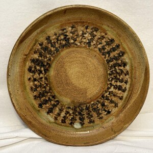May include: A round ceramic plate with a speckled tan and brown glaze. The center features a smooth, solid tan circle, surrounded by a textured ring of dark brown, leaf-like impressions. The outer rim is a smooth, light brown with subtle green tones.