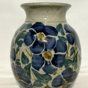 May include: A ceramic vase with a speckled off-white base and a hand-painted floral pattern featuring deep blue petals, yellow centers, and muted green leaves. The vase has a rounded body and a flared rim, showcasing a smooth, glazed finish.
