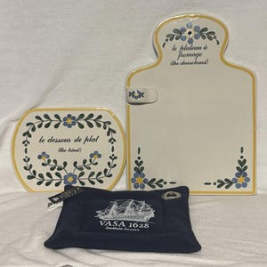 May include: A set of three ceramic serving pieces: a cheese board, a trivet, and a navy blue square with a ship graphic. The cheese board and trivet are white with yellow trim and floral accents. The cheese board has the text "le plateau à fromage (the cheese board)".
