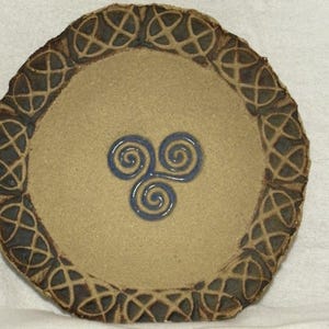 May include: A circular, tan ceramic plate with a dark brown Celtic knot border. The centre features a blue spiral design. The plate has a rustic, handmade appearance, with a slightly uneven edge.