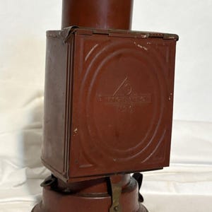 May include: A vintage, maroon-colored lantern with a rectangular body and a circular base. The lantern has a cylindrical top and a small handle. The front of the lantern has a circular design with the text "INGERSOLL" and a logo.