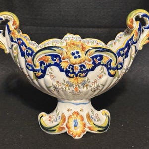 May include: A ceramic footed bowl featuring a cream-coloured base with intricate hand-painted floral and scroll motifs in shades of deep blue, vibrant yellow, and orange. The vessel has two ornate, curved handles and a scalloped rim edge.