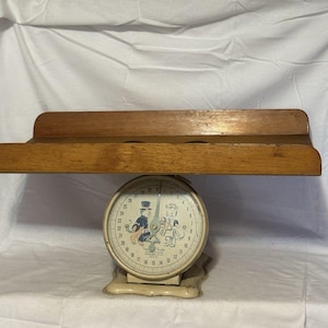 Könnte beinhalten: Vintage Küchenwaage mit einem Holzregal. Die Waage hat ein cremefarbenes Zifferblatt mit einer skurrilen Illustration eines Mannes und einer Frau. Die Waage misst bis zu 13,6 kg. Das Holzregal ist oben an der Waage befestigt.