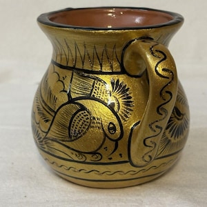 May include: A small, rounded ceramic mug with a metallic gold finish and black hand-painted details. The exterior features intricate line drawings of birds and floral patterns. The handle is also gold with black decorative lines, and the interior is a plain, unglazed terracotta color.
