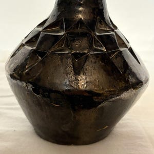 Op de afbeelding: Een donkerbruine, geglazuurde keramische vaas met een brede, ronde buik en een smalle hals. Het bovenste gedeelte van de vaas is voorzien van een herhalend geometrisch patroon van verzonken driehoeken, wat zorgt voor een gefacetteerd oppervlak.