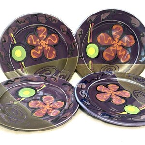 May include: A set of four round ceramic plates featuring a deep purple background with a large orange five-petaled flower, a small lime green circle, and abstract dark swirls. The lower portion of each plate has a muted bronze-colored band.