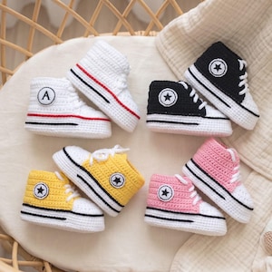 May include: A collection of crocheted baby sneakers in various colors, including white, black, yellow, and pink. Each shoe features a star emblem, laces, and a striped sole. The white sneakers have a red stripe and the letter "A".