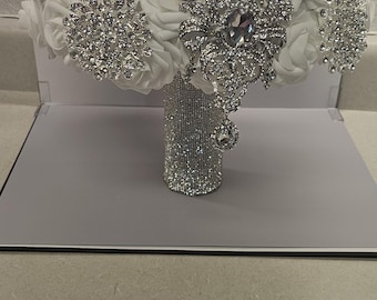 Puede incluir: Un ramo de novia con rosas de tela blancas adornadas con broches plateados brillantes y detalles de perlas. El mango está envuelto en pedrería brillante, creando un aspecto glamuroso y elegante.